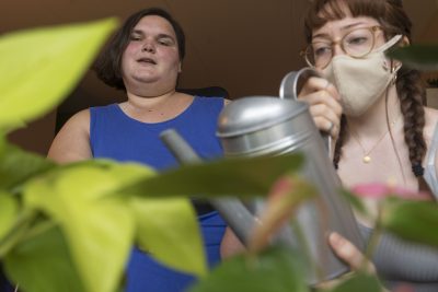 A close-up image framed by bright green leaves. Beyond the leaves, Marley is wearing a mask and wielding a silver watering can as Kennedy verbally directs her, maskless. 