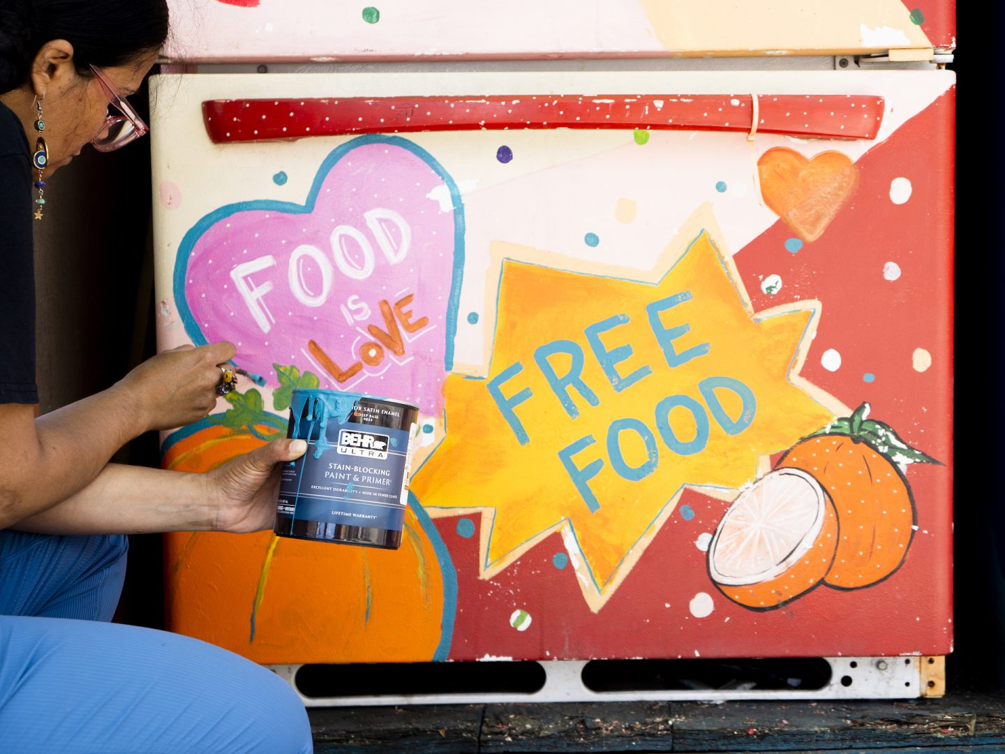Image features a person on the left painting the bottom of a refrigerator. A pink star with a blue outlines reads "Food is Love", and a orange star with a yellow outline reads "Free Food". There are oranges in the bottom right corner of the refrigerator.