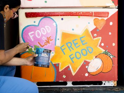 Image features a person on the left painting the bottom of a refrigerator. A pink star with a blue outlines reads 