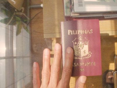 Image of a brown hand touching a burgundy passport with gold font that reads “Pilipinas Pasaporte” and features the Filipino crest in the center. The passport is sitting on a glass table- beneath it there are a stack of books sitting on wood floor.