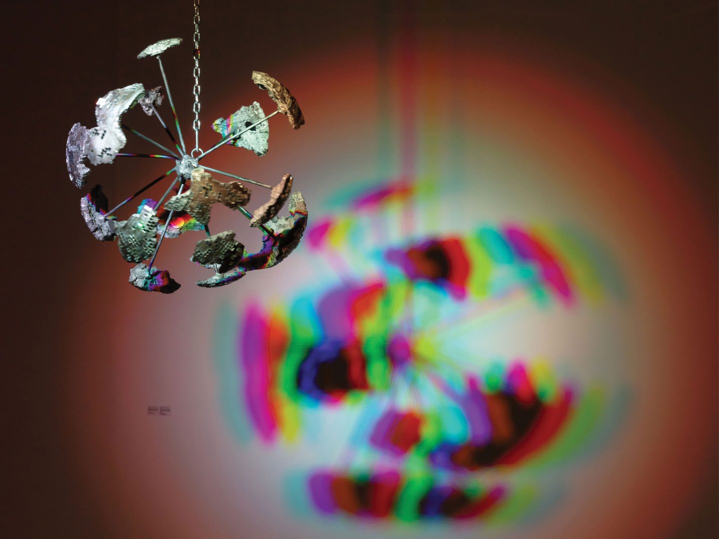 Suspended and fractured disco ball that is casting a rainbow like shadow against a wall.