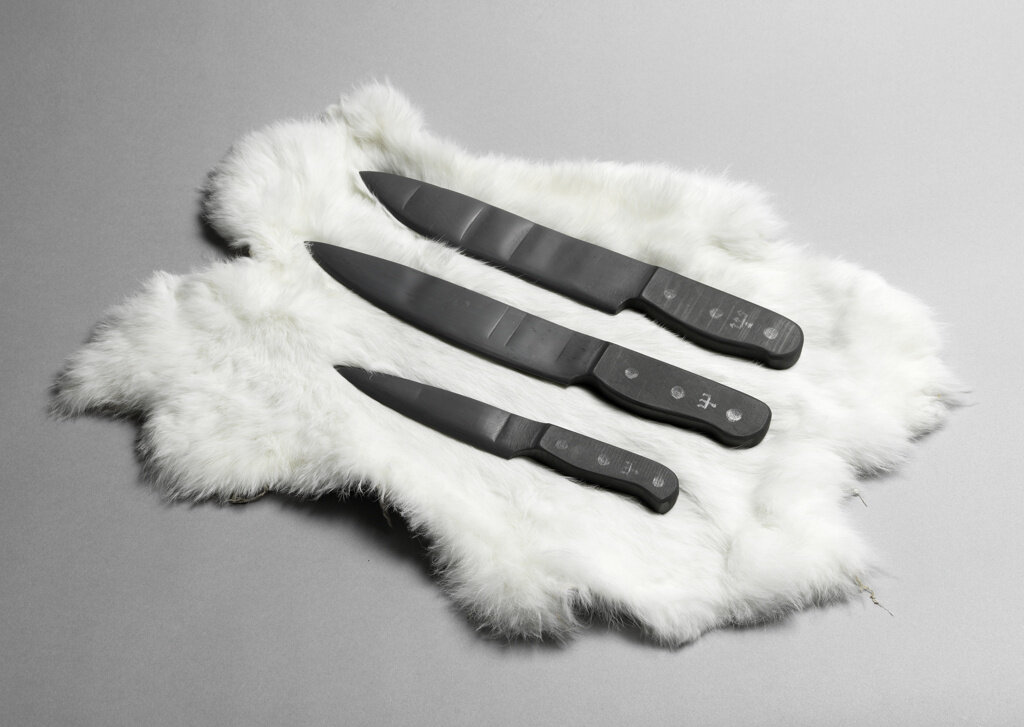Three black knives in various sizes resting on a white fur against a gray background.